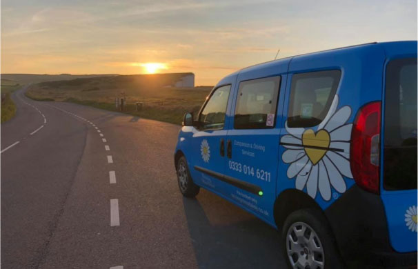 A blue driving Miss Daisy vehicle with a large decal of a white daisy with a yellow heart at its centre. Parked at the edge of the road, the sun is setting in the distance.