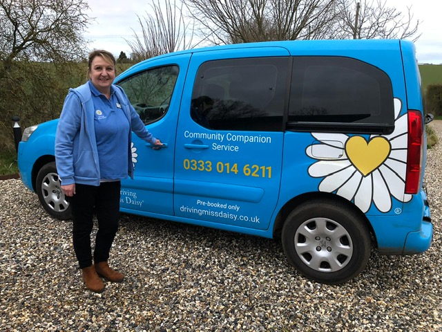 A female companion driver is standing next to a Driving Miss Daisy vehicle. The vehicle is blue with a large decal of a white daisy with a yellow heart at its centre.