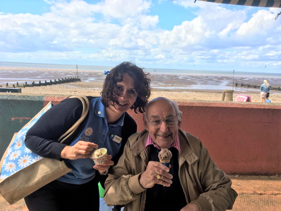 A woman in a driving miss daisy uniform with a friendly smile is at the beach with a gentleman in a wheelchair. Both are enjoying an ice cream.