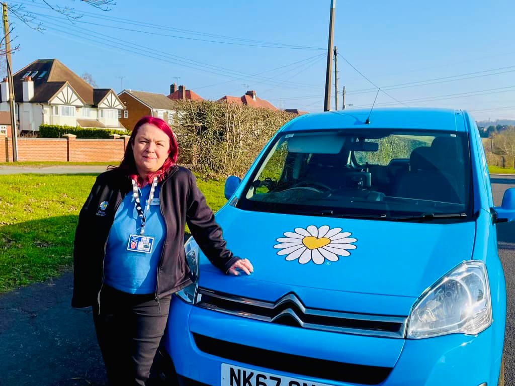 A redheaded women in a blue Driving Miss Daisy uniform, is smiling as she leans on a blue Driving Miss DAisy Car with a large decal of a daisy with a yellow heart at it's centre.